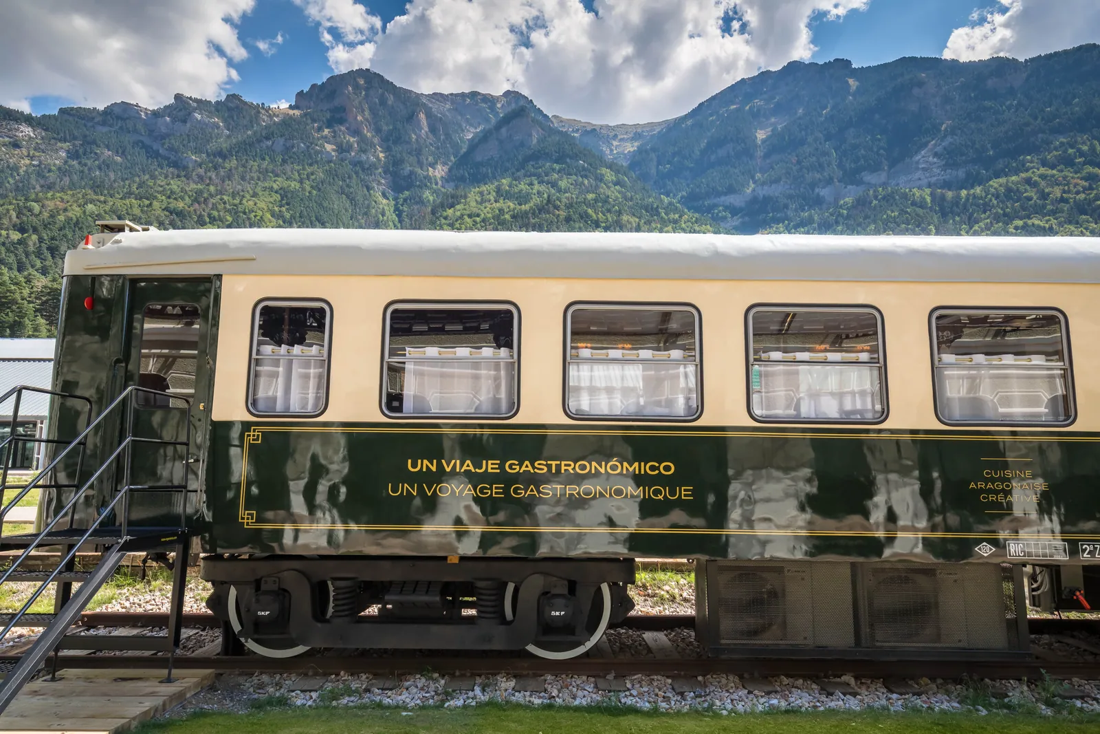 Un vagón de tren verde y crema, el restaurante Canfranc Express, sobre una vía frente a montañas verdes y rocosas bajo las nubes